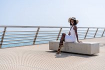 Вид сбоку триумфальная афроамериканка в стильном белом платье, сидящая со скрещенными ногами на каменной скамейке на городской набережной и наслаждающаяся видом на море в солнечную погоду — стоковое фото