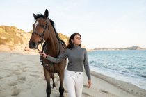 Young female walking with chestnut stallion looking away against wavy ocean — Stock Photo