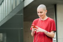 Jovem feliz homossexual fêmea em t-shirt e fones de ouvido com celular olhando para a tela enquanto ouve música — Fotografia de Stock
