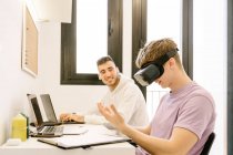 Side view of positive young male employee in casual clothes experiencing virtual reality in modern headset while working on laptop with smiling ethnic colleague — Photo de stock