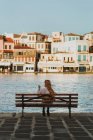 Vista posterior viajera femenina sin rostro descansando en el banco en el antiguo paseo marítimo del canal de la ciudad histórica en Chania Creta - foto de stock