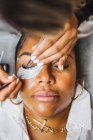 High angle of crop unrecognizable cosmetologist with tweezers applying fake eyelashes for extension on eye of ethnic client in salon — Stock Photo