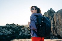 Vista lateral de la joven exploradora alegre en sudadera con capucha y gafas de sol sonriendo mientras admira la naturaleza durante el trekking en el valle montañoso rocoso en el día soleado - foto de stock