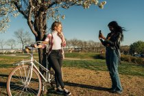 Vista lateral de la mujer negra tomando fotos de una amiga de pie con bicicleta cerca del árbol en el jardín de primavera - foto de stock