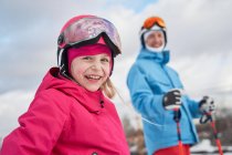 Веселая маленькая девочка в лыжном шлеме и активной одежде стоит рядом с размытым отцом в снежной зимней местности и смотрит в камеру с улыбающимся лицом — стоковое фото
