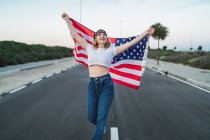 Entzückte Amerikanerin steht bei Sonnenuntergang mit nationaler US-Flagge auf der Fahrbahn und blickt nach oben — Stockfoto