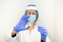 Médica em luvas de látex e proteção facial preenchendo seringa de frasco com vacina preparando-se para vacinar paciente na clínica durante surto de coronavírus — Fotografia de Stock