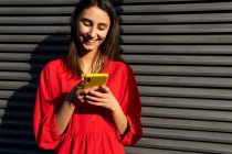 Giovane contenuto femminile in rosso usura chat sul telefono cellulare alla luce del sole su sfondo grigio — Foto stock