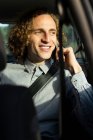 Cheerful young stylish haired male sitting on driver seat and speaking on mobile phone during trip through nature in sunny summer day — Photo de stock