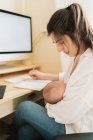 Mãe adulta amamentando recém-nascido com leite materno enquanto toma notas no caderno à mesa durante o dia — Fotografia de Stock