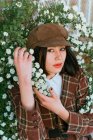 Young gentle ethnic female in checkered wear with beret touching blossoming plant while looking at camera on street — Fotografia de Stock