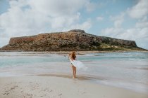 Повна довга самка в чорному купальнику стоїть на піщаному пляжі Балос проти скелястої скелі і дивиться у ясну сонячну погоду. — стокове фото