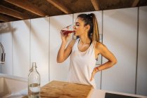 Female athlete in sportswear with hand on hip enjoying refreshing drink from glass at table with bottle of water — Foto stock