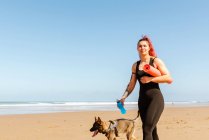 Atleta sonriente con alfombra enrollada y botella de agua paseando con perro de pura raza en la costa arenosa del mar mientras mira hacia otro lado - foto de stock