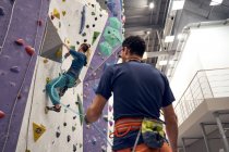 Desde abajo de valiente atleta escalando la pared artificial en el centro de bouldering bajo la supervisión de instructor profesional - foto de stock