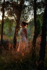 Полное тело спокойной женщины в белом платье стоя у ствола дерева в темных лесах в тихом закате света — стоковое фото