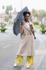 Full body young trendy African American female in warm coat standing with umbrella on modern city street and looking at camera — Stock Photo