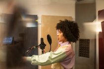 Black female singer performing song against microphone with pop filter while standing with hand on hip and looking forward in sound studio — Stock Photo