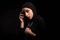 Привлекательная молодая исламская женщина в черной одежде и хиджабе нежно касается лица и смотрит в камеру. — стоковое фото