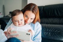 Молодая женщина делает селфи с очаровательным малышом на мобильном телефоне, проводя время дома — стоковое фото