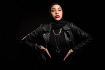 Attractive young Islamic female wearing black outfit with leather jacket and hijab gently looking at camera on black studio — Stock Photo