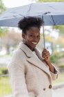 Young trendy African American female in warm coat standing with umbrella on modern city street and looking at camera — Stock Photo