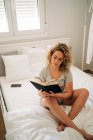 Сверху положительная молодая женщина с кудрявыми светлыми волосами в трусиках и очках улыбается, сидя на уютной кровати и читая интересную книгу — стоковое фото