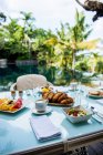 From above of comfortable wicker armchair with soft seats and cushions placed near table served with various appetizing pastries and fruits during breakfast in tropical resort - foto de stock