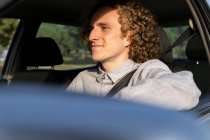 Happy young haired male looking away through open window of car while sitting at driver seat — Photo de stock