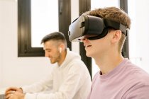 Side view of positive young male employee in casual clothes experiencing virtual reality in modern headset while working on laptop with smiling ethnic colleague — Foto stock
