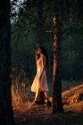 Full body of calm female in white dress standing at tree trunk in dark woods in calm sundown light — Foto stock