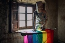 Pessoa irreconhecível em traje de prata e caixa na cabeça engomar bandeira LGBTQ enquanto está em pé em quarto abandonado miserável — Fotografia de Stock