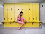 Side view of female swimmer in swimwear with wet hair and towel sitting on bench against yellow lockers and looking down — Stock Photo