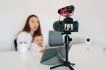 Розмита молода мама з милою дитиною на колінах говорить і записує відео на камеру для особистого блогу, сидячи за столом вдома — стокове фото