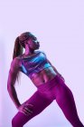 Jeune athlète afro-américaine musclée en vêtements de sport avec des tresses afro et les mains sur les hanches regardant loin dans la lumière néon — Photo de stock