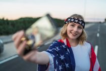 Mulher americana Patriótica envolta em bandeira nacional dos EUA tomando selfie no telefone celular enquanto estava de pé na estrada — Fotografia de Stock