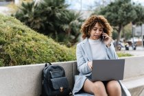 Fokussierte junge afrikanisch-amerikanische Frau in blauem Mantel arbeitet am Netbook und telefoniert am Handy, während sie an klaren Frühlingstagen auf einer steinernen Bank im Stadtpark sitzt — Stockfoto
