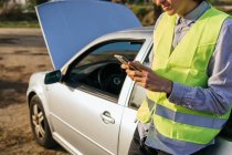 Crop unrecognizable male driver in green safety vest using mobile phone and asking for help after car accident on country road — Stock Photo