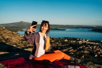Side view of happy young female hiker in casual clothes eating sandwich and taking selfie with mobile phone while sitting on blanket on rocky hill over sea against cloudless blue sky — Stock Photo