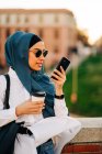 Ethnic female in headscarf and stylish sunglasses standing with takeaway drink on street and recording voice message on mobile phone — Foto stock