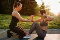 Side view of slim female athlete doing abdominal crunches with help of sportswoman during training at sunset in park — Stock Photo