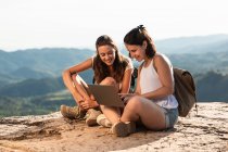Entzückte reisende Freundinnen, die auf Felsen im Hochland sitzen und sich beim Sommertrekking Filme auf Netbook ansehen — Stockfoto