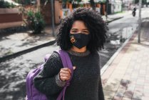 Side view of delighted ethnic female student with Afro hairstyle and backpack standing on street on sunny day with face mask and looking at camera - foto de stock