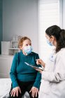 Female medic in uniform with tablet speaking with senior woman in sterile mask on consultation while looking at each other during coronavirus pandemic — Stock Photo