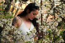 Junge Frau mit tätowiertem Arm trägt weißes Kleid und steht in Blumen des Baumes und blickt nach unten — Stockfoto