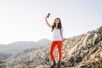 Pieno corpo di positivo giovane viaggiatore femminile con i capelli ricci scuri in abiti casual sorridente mentre prende selfie sul telefono cellulare durante il trekking in montagna nella giornata di sole — Foto stock
