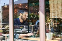 Through glass view of adult ethnic male remote employee working on netbook at table — Stock Photo