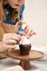 Cultivo joven hembra decoración cupcake de chocolate con crema batida dulce y flores en pie de pastel mientras se cocina en casa - foto de stock
