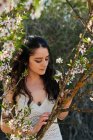 Young female with tattooed arm wearing white dress and standing in flowers of tree looking down — Photo de stock