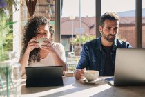 Ethnic female remote employee enjoying hot drink against tablet and male partner with laptop at table in sunlight — Stock Photo